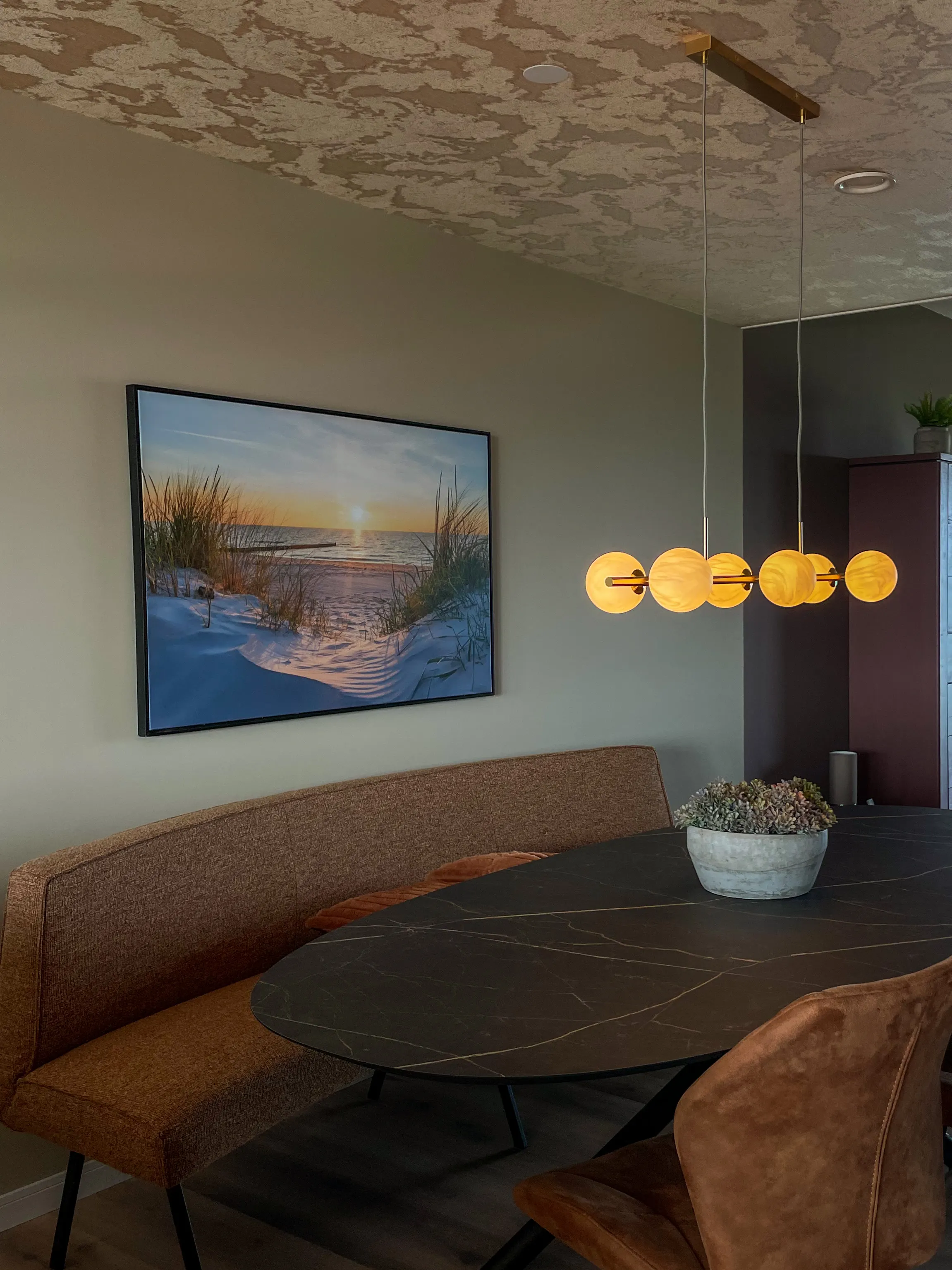 Eetbank en ronde tafel onder een schilderij van de duinen, met een bollamp erboven